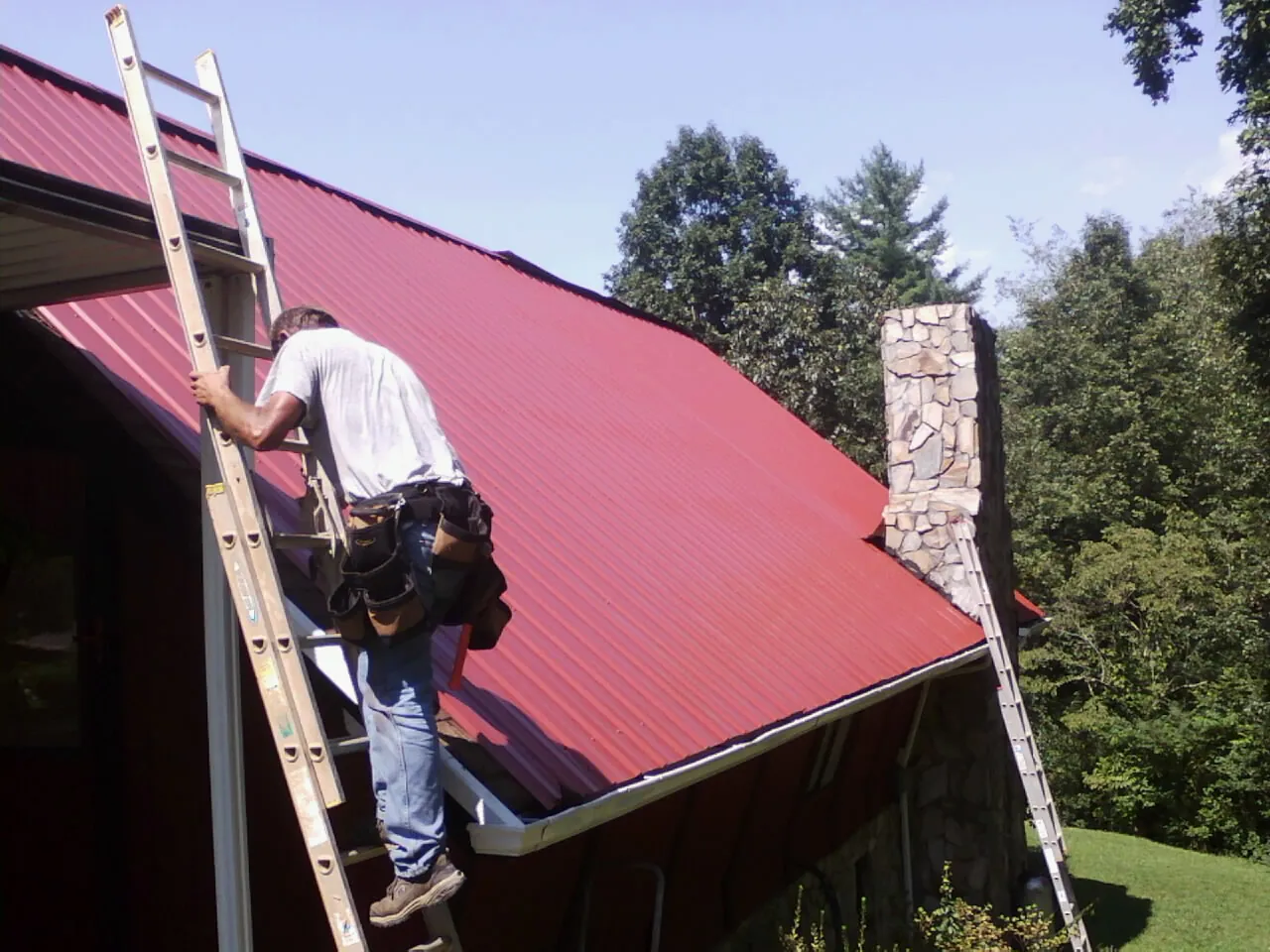 Roofer on a ladder beside a red metal roof and stone chimney — Soffit & Fascia Repair by Roofing Brothers in Smoaks